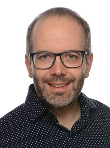 Herr Kaiser mit Brille, Hemd mit Punkten und kurzem Bart, frontal.